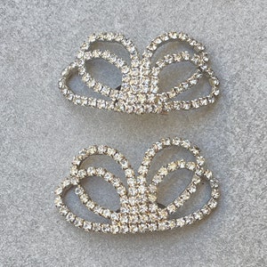 May include: Two rhinestone shoe clips in a bow shape. The clips are silver-toned and encrusted with clear rhinestones. The design features multiple loops and curves, creating a sparkling accessory.