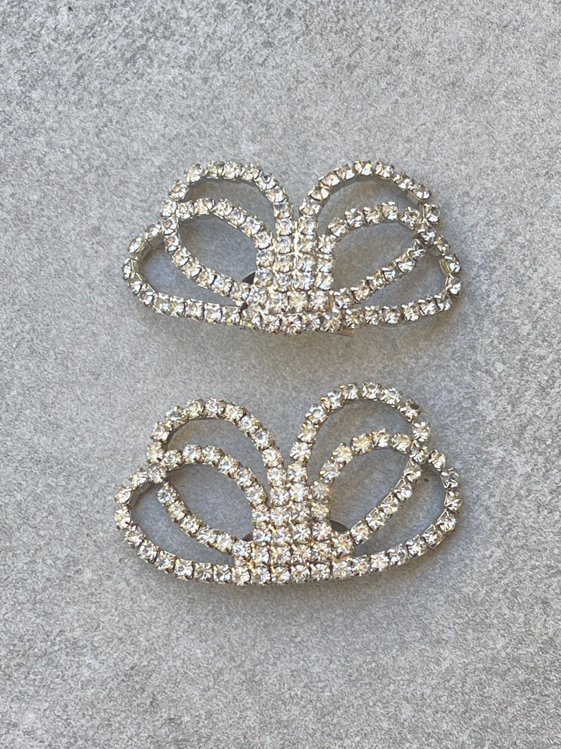 May include: Two rhinestone shoe clips in a bow shape. The clips are silver-toned and encrusted with clear rhinestones. The design features multiple loops and curves, creating a sparkling accessory.