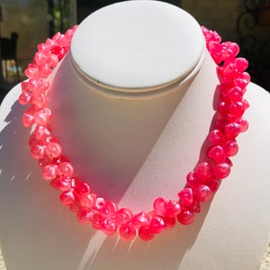 May include: A pink shell necklace with a gradient of color from light pink to dark pink. The necklace is made of small, round shells that are strung together on a thin cord.