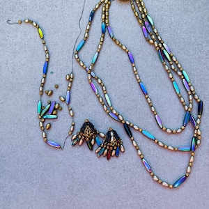 May include: A multi-strand necklace with iridescent beads and a gold clasp. The necklace is made of three strands of small, round, white beads and one strand of iridescent, oval-shaped beads. The clasp is a gold circle with a decorative design.