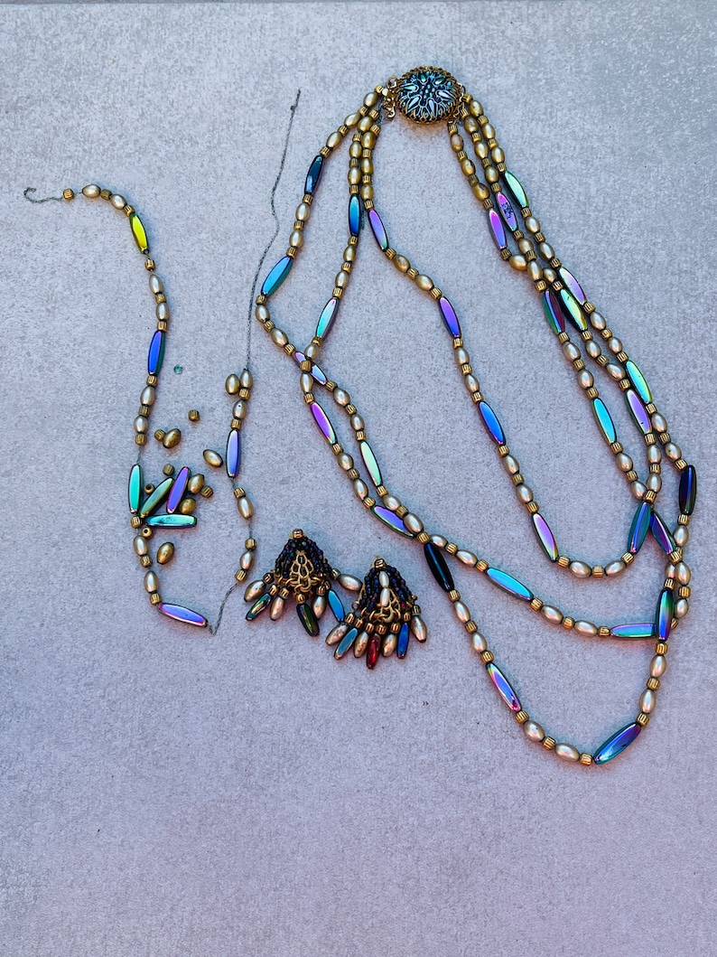 May include: A multi-strand necklace with iridescent beads and a gold clasp. The necklace is made of three strands of small, round, white beads and one strand of iridescent, oval-shaped beads. The clasp is a gold circle with a decorative design.