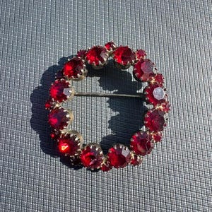 Puede incluir: Broche circular vintage con un anillo de gemas rojas brillantes. El broche tiene un engaste de metal plateado y un cierre de alfiler. Las gemas están facetadas, captando la luz y creando un efecto brillante. Una pieza de joyería clásica.