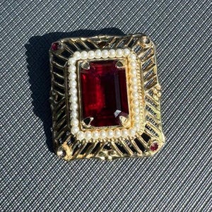 Pode incluir: Broche retangular dourado com uma grande pedra preciosa retangular vermelho escuro no centro. A pedra preciosa é cercada por uma fileira de pequenas pérolas brancas e uma moldura de filigrana dourada. Pequenas pedras vermelhas nos cantos.