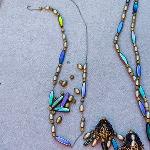 May include: Two strands of iridescent bead necklaces with gold accents and pearl-like beads. The necklaces are partially strung and have loose beads scattered around them. The necklaces have decorative tassels with iridescent beads and gold accents.