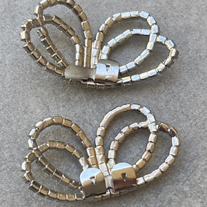 May include: Two silver-toned, butterfly-shaped shoe clips. Each clip is adorned with numerous small, square, clear rhinestones. The clips have a metal clasp on the back for attachment to shoes or other accessories.