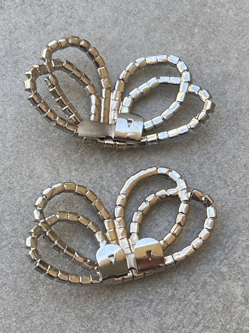 May include: Two silver-toned, butterfly-shaped shoe clips. Each clip is adorned with numerous small, square, clear rhinestones. The clips have a metal clasp on the back for attachment to shoes or other accessories.