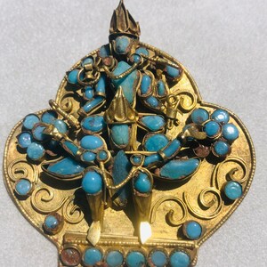 May include: A gold pendant with a turquoise blue figure in the center. The figure is surrounded by intricate gold designs and turquoise blue stones.