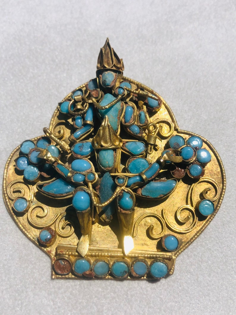 May include: A gold pendant with a turquoise blue figure in the center. The figure is surrounded by intricate gold designs and turquoise blue stones.