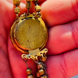May include: A gold-toned necklace with a round pendant featuring a detailed design. The pendant has a clasp on the back and is attached to a chain with multiple strands of beads.