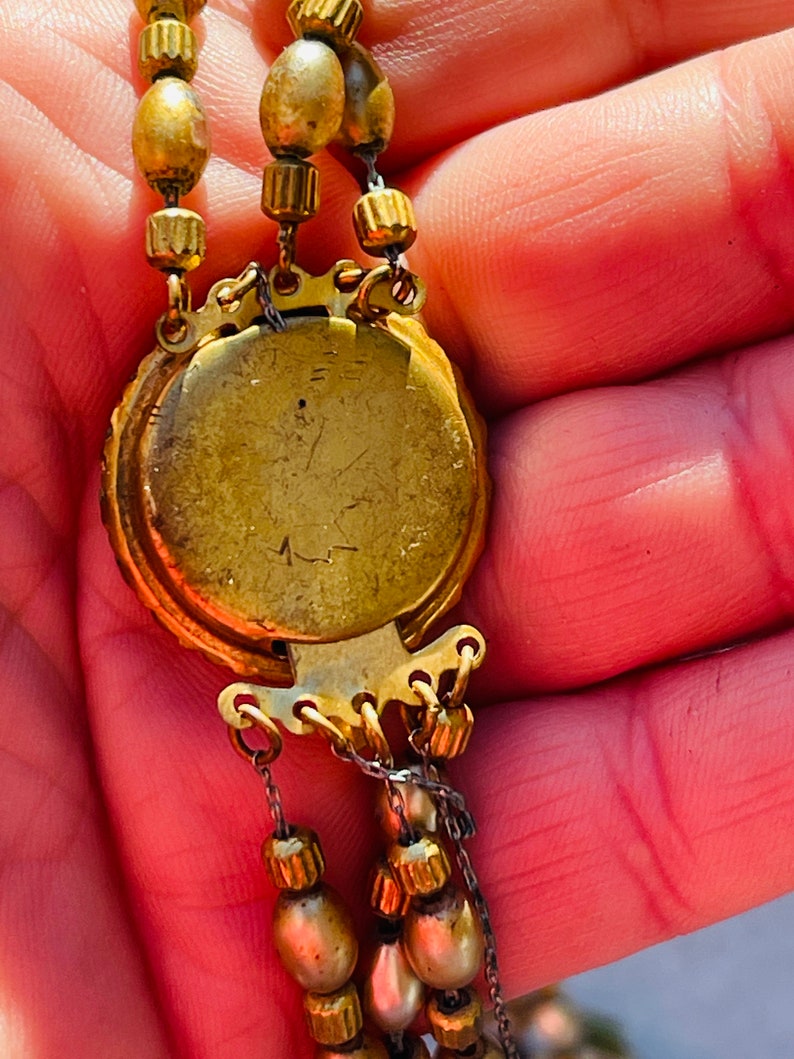 May include: A gold-toned necklace with a round pendant featuring a detailed design. The pendant has a clasp on the back and is attached to a chain with multiple strands of beads.