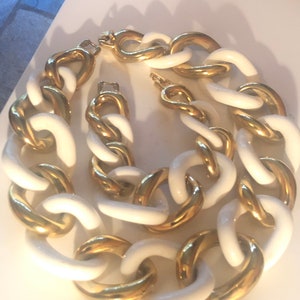 Puede incluir: Un conjunto de collar y pulsera de cadena blanco y dorado. Los eslabones son grandes y gruesos, con un exterior de cerámica blanca y un interior de metal dorado.