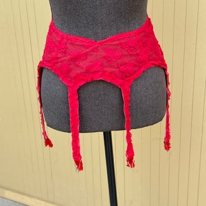 Puede incluir: Una liga de encaje rojo con motivos florales. El cinturón tiene cuatro ligueros con clips metálicos. La prenda se exhibe sobre un torso de maniquí gris sobre un fondo amarillo y blanco.