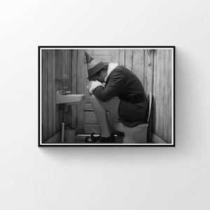 May include: Black and white framed print of a person dressed as an elf, sitting on a toilet in a small bathroom. The elf is wearing a green and red hat, a red jacket with white trim, and black shoes.