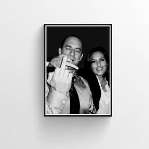 May include: Black and white framed photograph of two people. One person is making a rude hand gesture, while the other person is smiling. The photograph is displayed on a white wall.