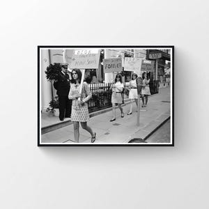 May include: Black and white photograph of a protest. Several women in dresses are holding signs that read "Mini Skirts Forever" and other slogans. A police officer stands nearby. The scene is on a sidewalk in front of a building.