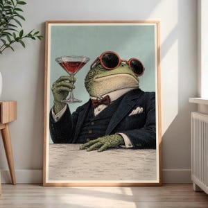 May include: A framed wall art print featuring an anthropomorphic toad wearing a dark blue suit, a white collared shirt, a red bow tie, and round red-rimmed sunglasses. The toad holds a stemmed glass filled with a red liquid against a light blue background.