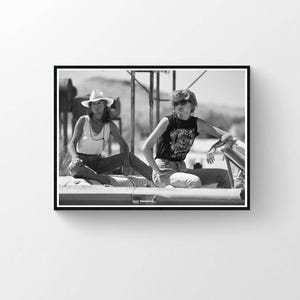 Puede incluir: Fotografía en blanco y negro de dos personas sentadas en el borde de un coche descapotable. Una persona lleva una camiseta sin mangas blanca, un sombrero y vaqueros. La otra persona lleva una camiseta negra con gráficos, gafas de sol y vaqueros. El fondo es un paisaje desértico.