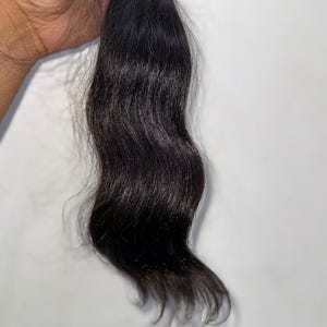 Puede incluir: Un mechón de extensiones de cabello humano castaño oscuro, sujetas con una banda amarilla. El cabello tiene una textura ondulada y un color natural profundo. El cabello se muestra sobre un fondo blanco liso.