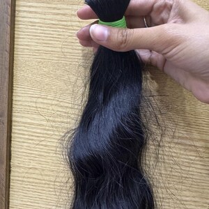 Puede incluir: Un mechón de extensiones de cabello negro oscuro, sujetas con una banda verde. El cabello es ondulado y parece natural, con un ligero brillo. El fondo es una superficie de madera de color claro.