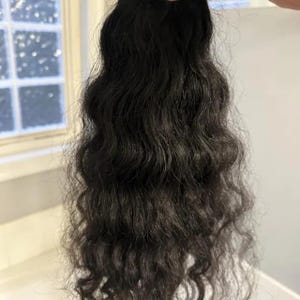 Puede incluir: Un mechón de cabello oscuro y ondulado. El cabello es de color negro intenso y tiene un aspecto natural y texturizado. Las hebras son gruesas y llenas, con un ligero rizo. El cabello se mantiene unido en la parte superior, lo que sugiere extensiones o una peluca.