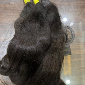 May include: Three bundles of dark brown human hair extensions, each secured at the top with a bright yellow elastic band. The hair has a natural wavy texture and a glossy finish, with the strands flowing downward from the tied sections.
