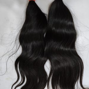 May include: Two bundles of dark, wavy hair, each secured with a coral-colored band at the top. The hair is long and flowing, with a natural wave pattern. The background is a plain, light surface.