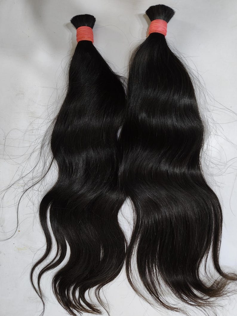 May include: Two bundles of dark, wavy hair, each secured with a coral-colored band at the top. The hair is long and flowing, with a natural wave pattern. The background is a plain, light surface.