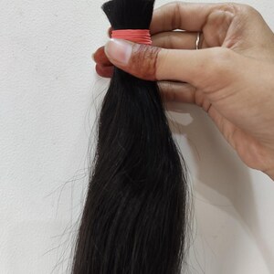 May include: A bundle of dark black hair, held together with a pink rubber band. The hair is long and straight, with a slight sheen. The hair is being held by a hand.