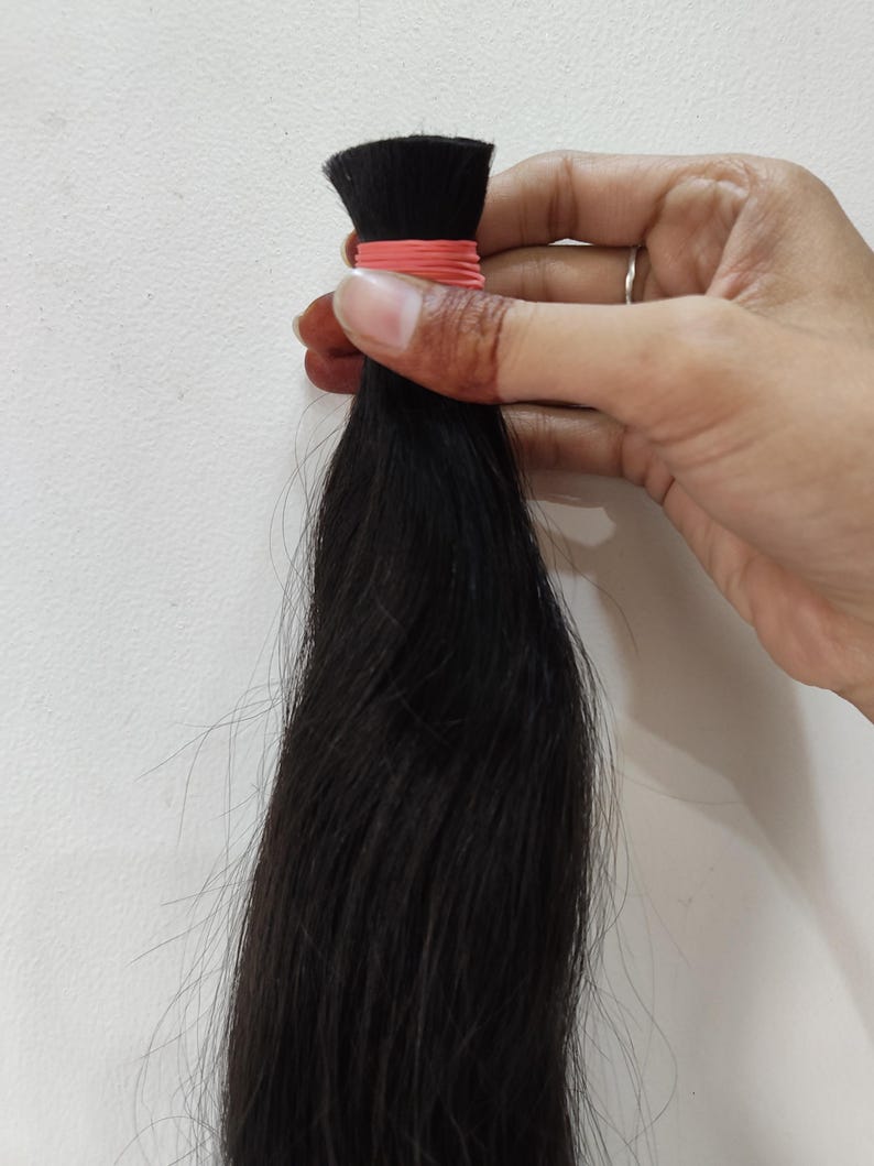 May include: A bundle of dark black hair, held together with a pink rubber band. The hair is long and straight, with a slight sheen. The hair is being held by a hand.