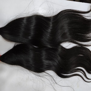 May include: Two bundles of dark, wavy hair are displayed against a light background. Each bundle is secured with a bright orange elastic band at the base. The hair appears to be long and flowing, with a natural sheen.