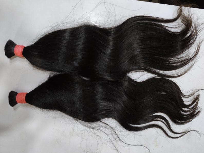 May include: Two bundles of dark, wavy hair are displayed against a light background. Each bundle is secured with a bright orange elastic band at the base. The hair appears to be long and flowing, with a natural sheen.