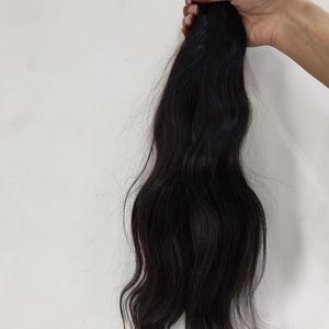 May include: A bundle of dark, wavy hair extensions held against a white background. The hair is a deep black color, with visible waves and a natural texture. The ends are slightly lighter, and the hair is tied with red bands.