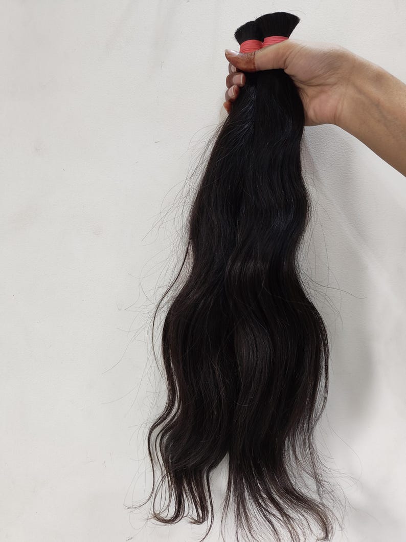 May include: A bundle of dark, wavy hair extensions held against a white background. The hair is a deep black color, with visible waves and a natural texture. The ends are slightly lighter, and the hair is tied with red bands.