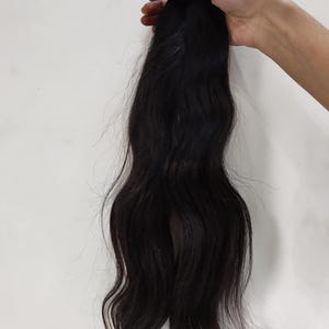 May include: A bundle of dark black hair extensions, held together with a pink band. The hair is wavy and appears to be natural, with a slight sheen. The background is a plain white surface.
