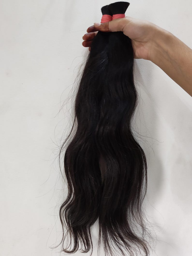 May include: A bundle of dark black hair extensions, held together with a pink band. The hair is wavy and appears to be natural, with a slight sheen. The background is a plain white surface.