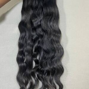 Puede incluir: Un paquete de extensiones de cabello ondulado negro oscuro. El cabello se mantiene unido en la parte superior, con los mechones ondulados que caen hacia abajo. El cabello parece natural y sin procesar, con una textura suave y fluida.