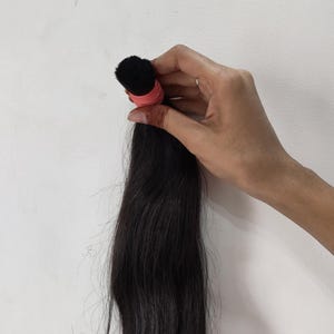 May include: A bundle of long, dark black hair, held together with a pink elastic band. The hair appears to be straight and sleek, with a natural shine. The hair is held against a plain white background.