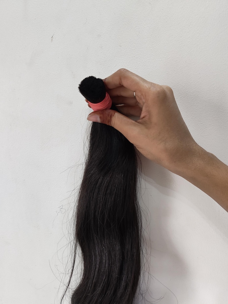 May include: A bundle of long, dark black hair, held together with a pink elastic band. The hair appears to be straight and sleek, with a natural shine. The hair is held against a plain white background.