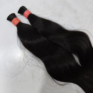 May include: Two bundles of black hair extensions. Each bundle is tied with a light orange band near the top. The hair is sleek and straight, with a natural shine. The background is a plain, light surface.