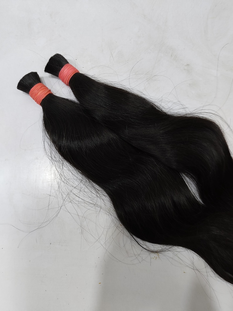 May include: Two bundles of black hair extensions. Each bundle is tied with a light orange band near the top. The hair is sleek and straight, with a natural shine. The background is a plain, light surface.