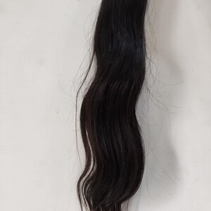 May include: A bundle of dark black hair extensions, wavy in texture, against a white background. The hair appears to be natural and unprocessed, with a slight sheen.
