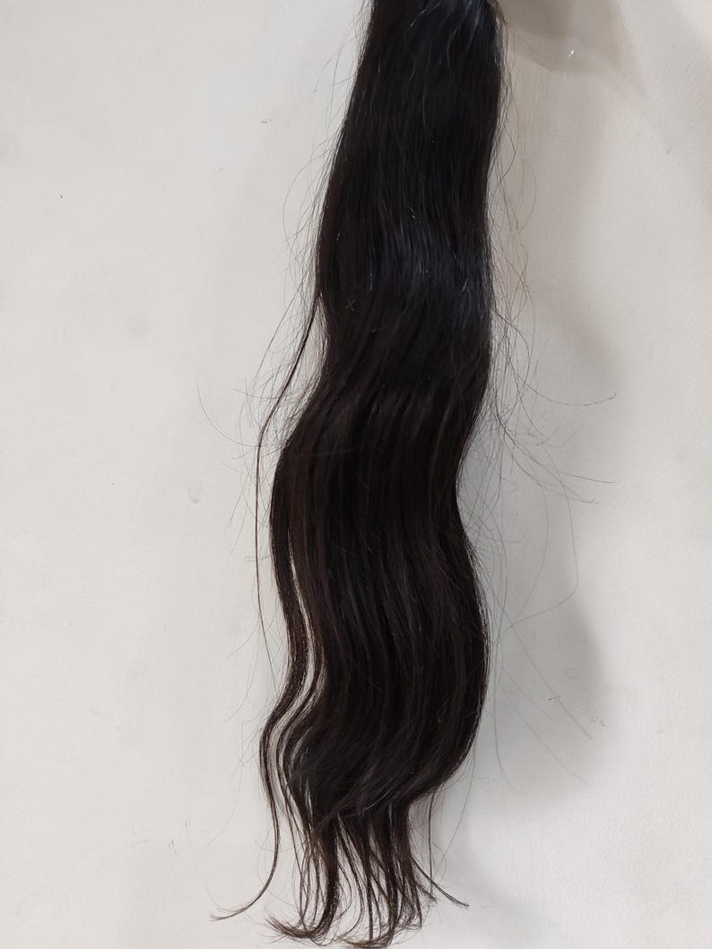 May include: A bundle of dark black hair extensions, wavy in texture, against a white background. The hair appears to be natural and unprocessed, with a slight sheen.