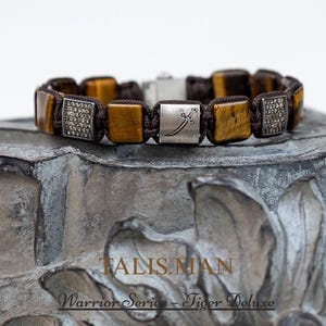 Op de afbeelding: Een bruine en gouden kralenarmband met vierkante tijgeroogstenen en zilveren accenten. De armband ligt op een grijs stenen oppervlak. De tekst "TALISMAN Warrior Series - Tiger Deluxe" is zichtbaar.