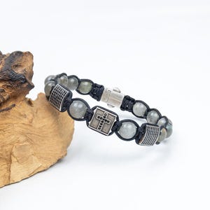 May include: A black cord bracelet with gray gemstone beads and silver-tone accents. The bracelet features a cross design and rectangular silver-tone pieces with black detailing. The clasp is silver-tone.