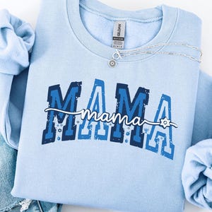 May include: A light blue sweatshirt with the word "MAMA" in navy and light blue block letters. The word "mama" is written in cursive underneath. A silver necklace and distressed blue jeans are next to the sweatshirt.