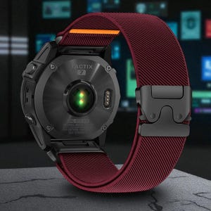 Puede incluir: Reloj inteligente negro con una correa de tela burdeos y un cierre negro. La esfera del reloj muestra el texto "TACTIX 7". La correa tiene un acento naranja. El reloj está diseñado para actividades al aire libre.