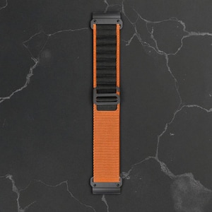 Puede incluir: Una correa de reloj naranja y negra con una correa de nailon tejido. La correa tiene una hebilla negra y conectores negros en cada extremo. La correa del reloj se muestra sobre una superficie gris oscuro.