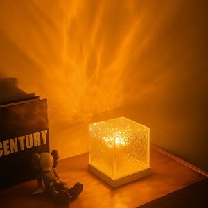 Puede incluir: Una lámpara de mesa con forma de cubo con un exterior de vidrio texturizado, que emite un brillo amarillo cálido. La lámpara se asienta sobre una base blanca, proyectando patrones de luz en la pared naranja. Un libro negro con la palabra "CENTURY" es visible.