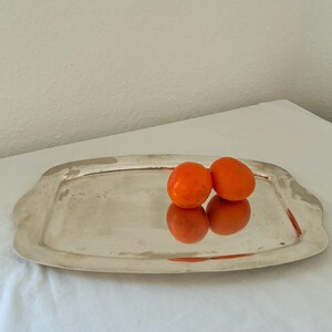 Puede incluir: Una bandeja rectangular de plata con bordes suavemente curvados. Dos caquis de color naranja brillante descansan sobre la bandeja, reflejándose en su superficie pulida. La bandeja está sobre una superficie blanca.