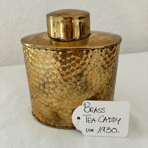 Puede incluir: Una tetería de latón dorado con textura martillada y tapa. La tetería tiene forma cilíndrica. Una etiqueta adjunta a la tetería dice "Brass Tea Caddy um 1930."